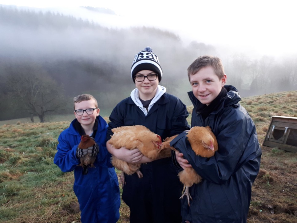 Three young people dressed in winter clothing holding a chicken each. It is a foggy winter day behind them.