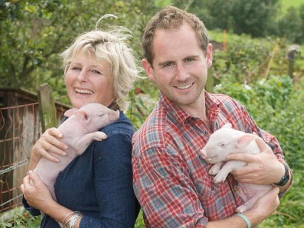 Jamie and Tish feilden stand back-to-back, smiling at the camera, while holding a piglet each
