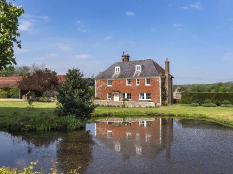 The beautiful Lewes farm house on a sunny day. The house is set behind a large pond, with trees to the left hand side.