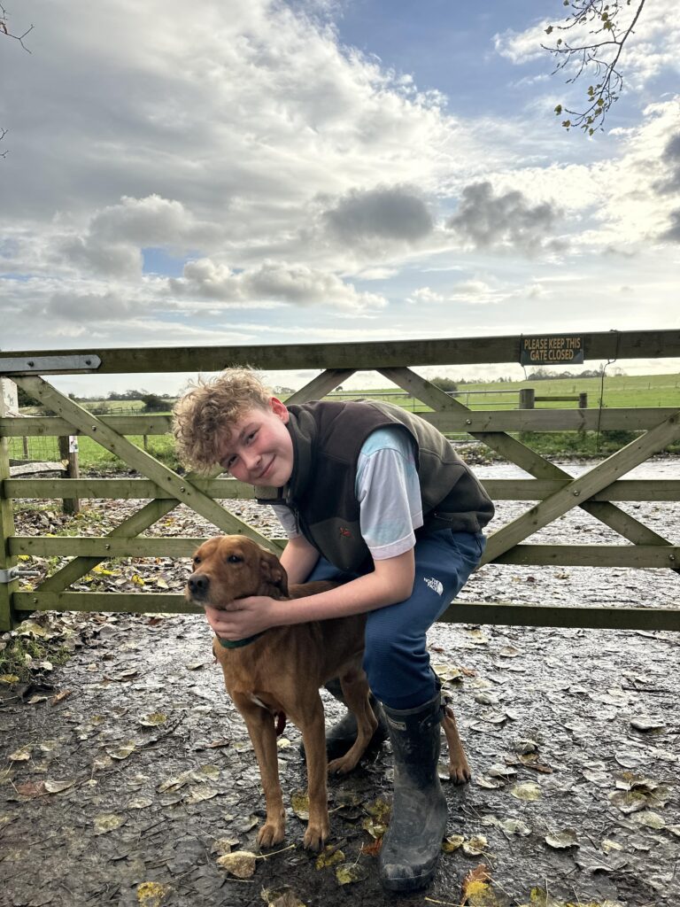 Riley petting a dog, standing outside in front of a farm gate with a blue sky and a field behind. 