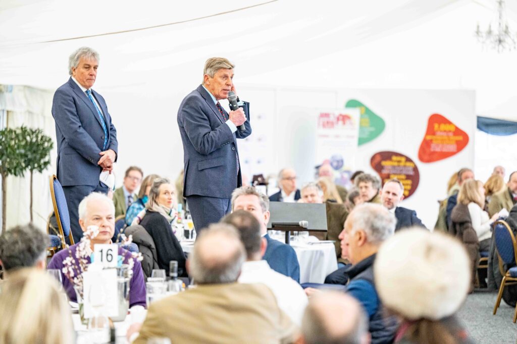 Brough Scott MBE speaking in front of a crowd of people sitting at tables