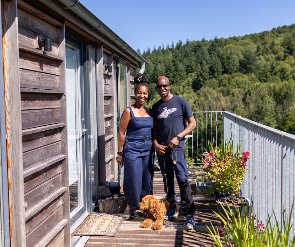 Michelle and her partner Herbie stood outside her flat at Jamie's Farm Monmouth, with a beautiful view of the woodlands behind them and their goldendoodle Dog, Bailey, at their feet 