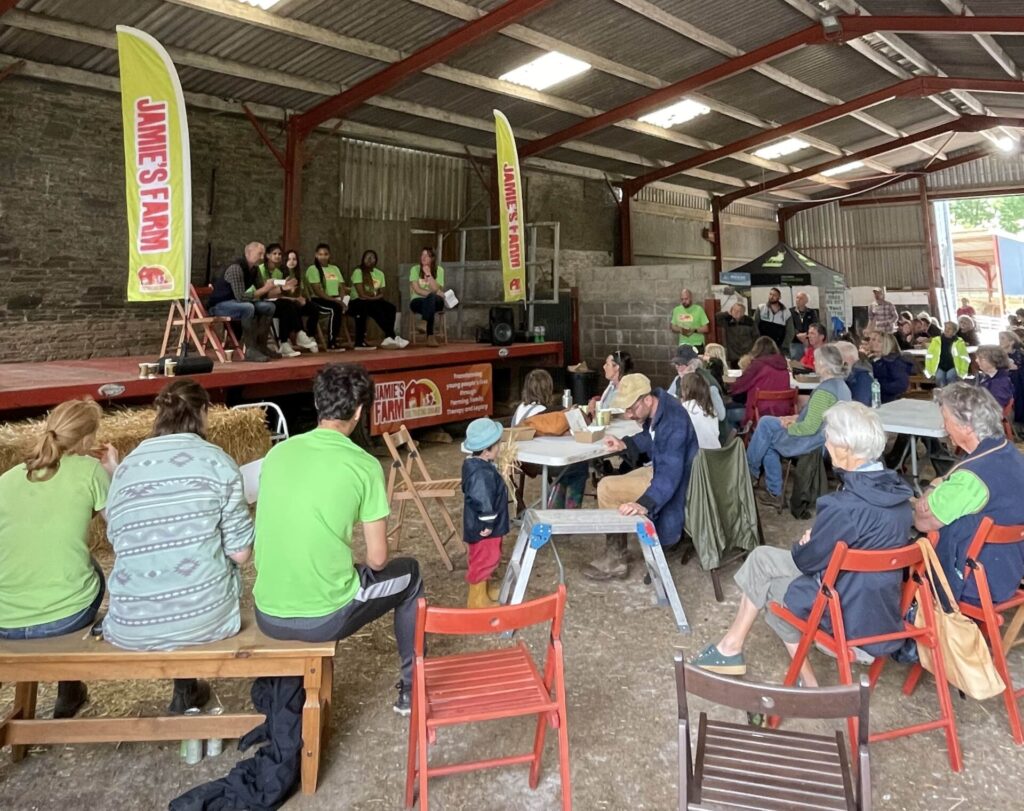 Group of people sitting at tables listening to six people talking inside a farm barn