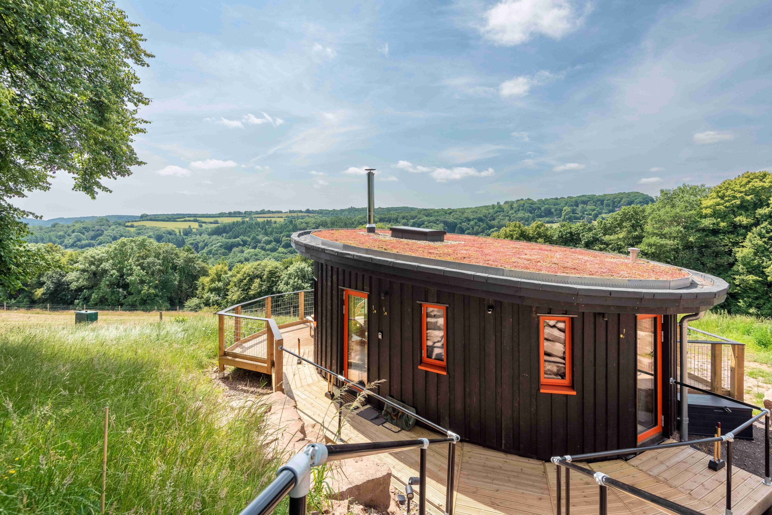 A wooden log cabin with red framed windows sat on a hillside with forests and hills on the horizon behind.