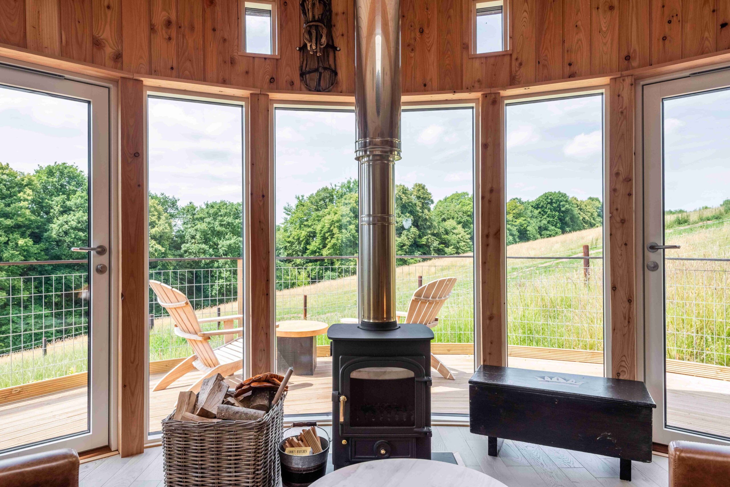 The inside of one of the cabins showing a log burner infront of panoramic windows with a view of hills and tress beyond.