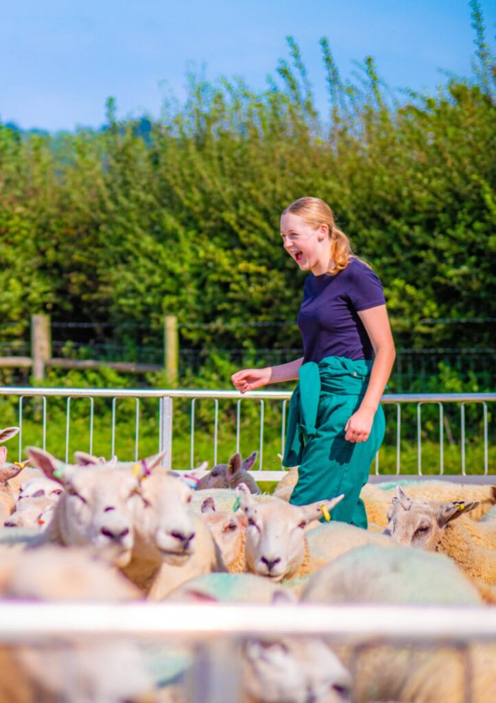 A girl in green overalls standing amongst a heard of sheep laughing joyfully