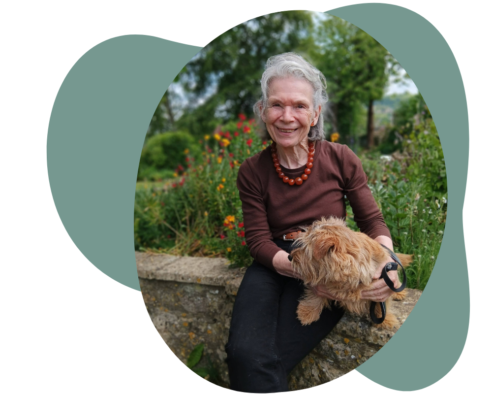 A photo of Sue smiling sitting on a wall in the garden at Hill House Farm. She is holding her dog on her lap with trees and flowers behind. 