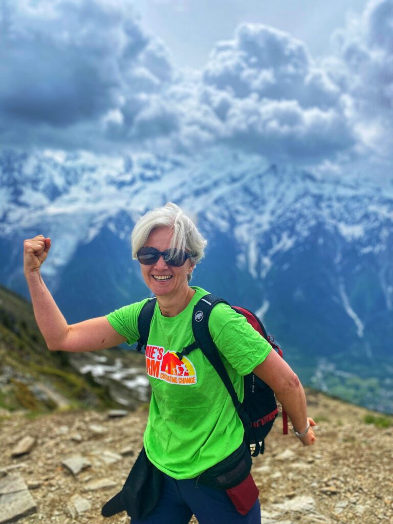 A lady posing with arms outstretched in front of alpine mountains wearing a Jamie's Farm green t-shirt. 