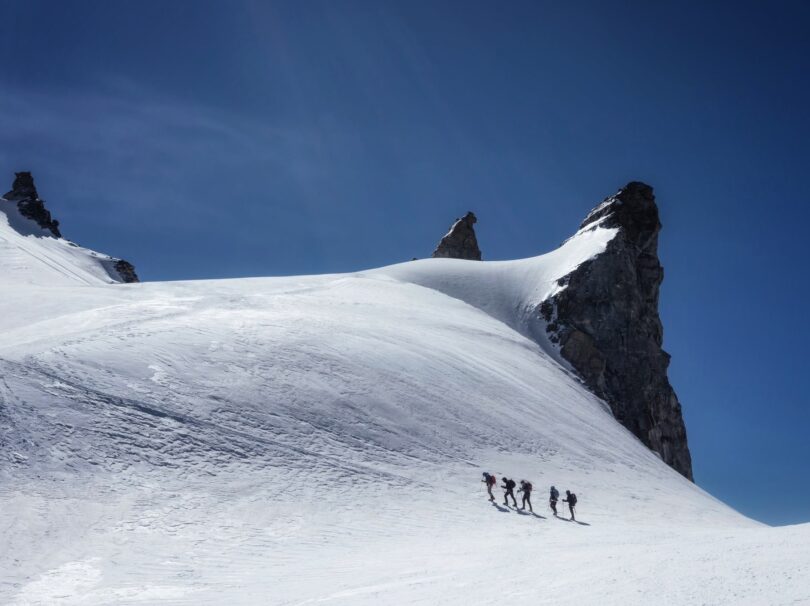 A group of climbers roped together ascending a snow-covered slope beneath jagged rock spires under a clear blue sky in an alpine environment.