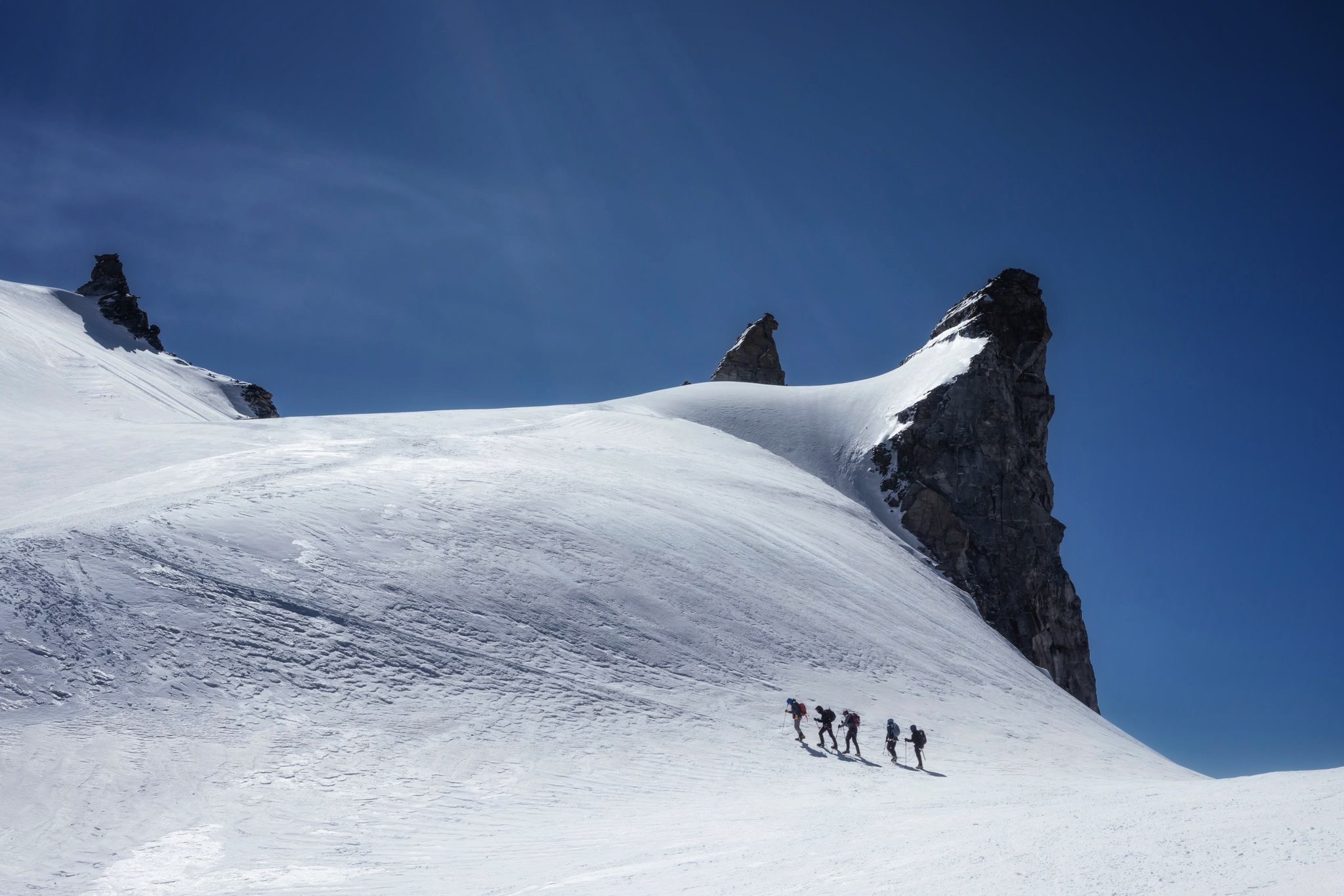 Walkers trekking in the distance at high altitude through the snow, in the sunshine.