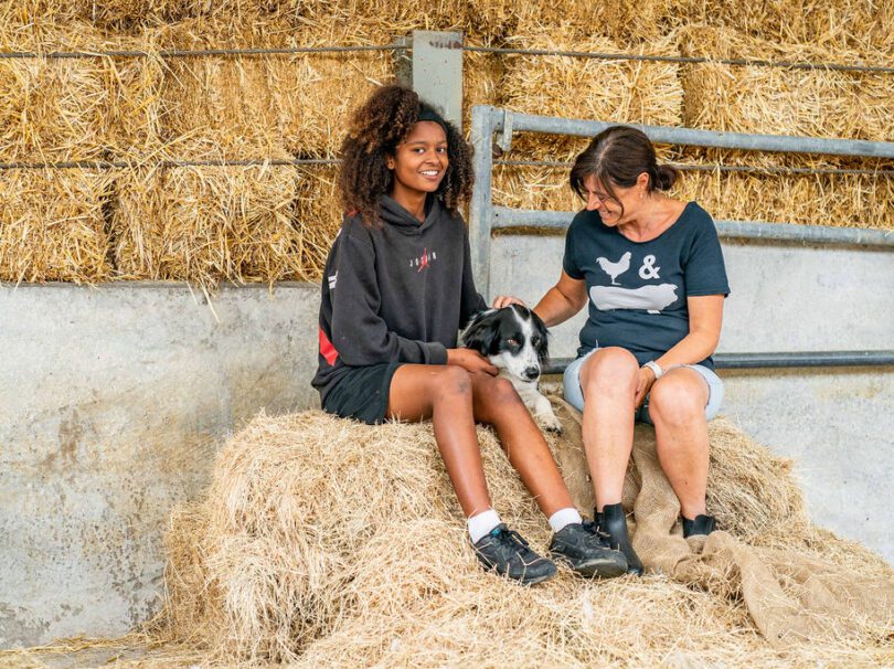 Girl and adult sitting on a hay bail with a dog between them