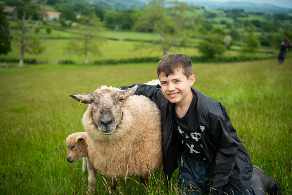 A boy crouching down in a field with his arm around a ewe with its lamb nearby.