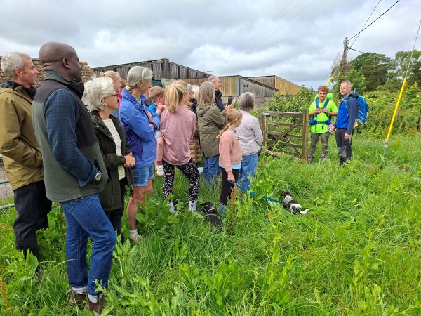 A crowd gathered in a field looking at two people wearing walking gear, getting ready to set off on a long journey. The sky is cloudy.
